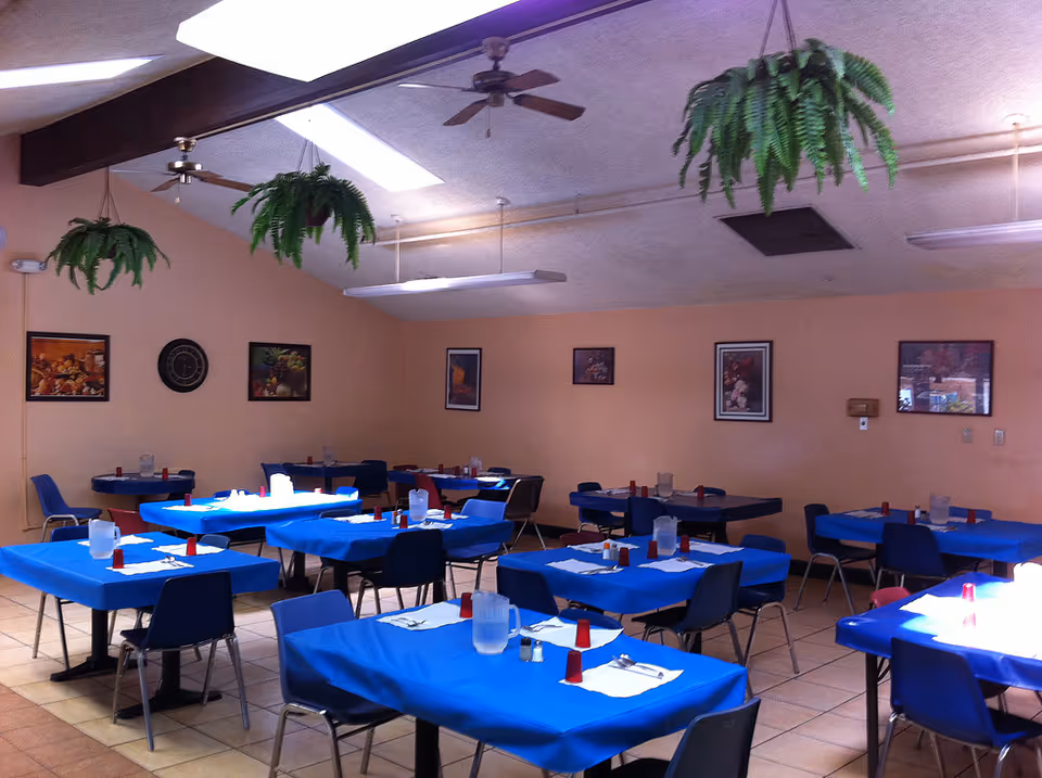Dining hall with blue tablecloths, many tables, chairs, pitchers, and wall art.