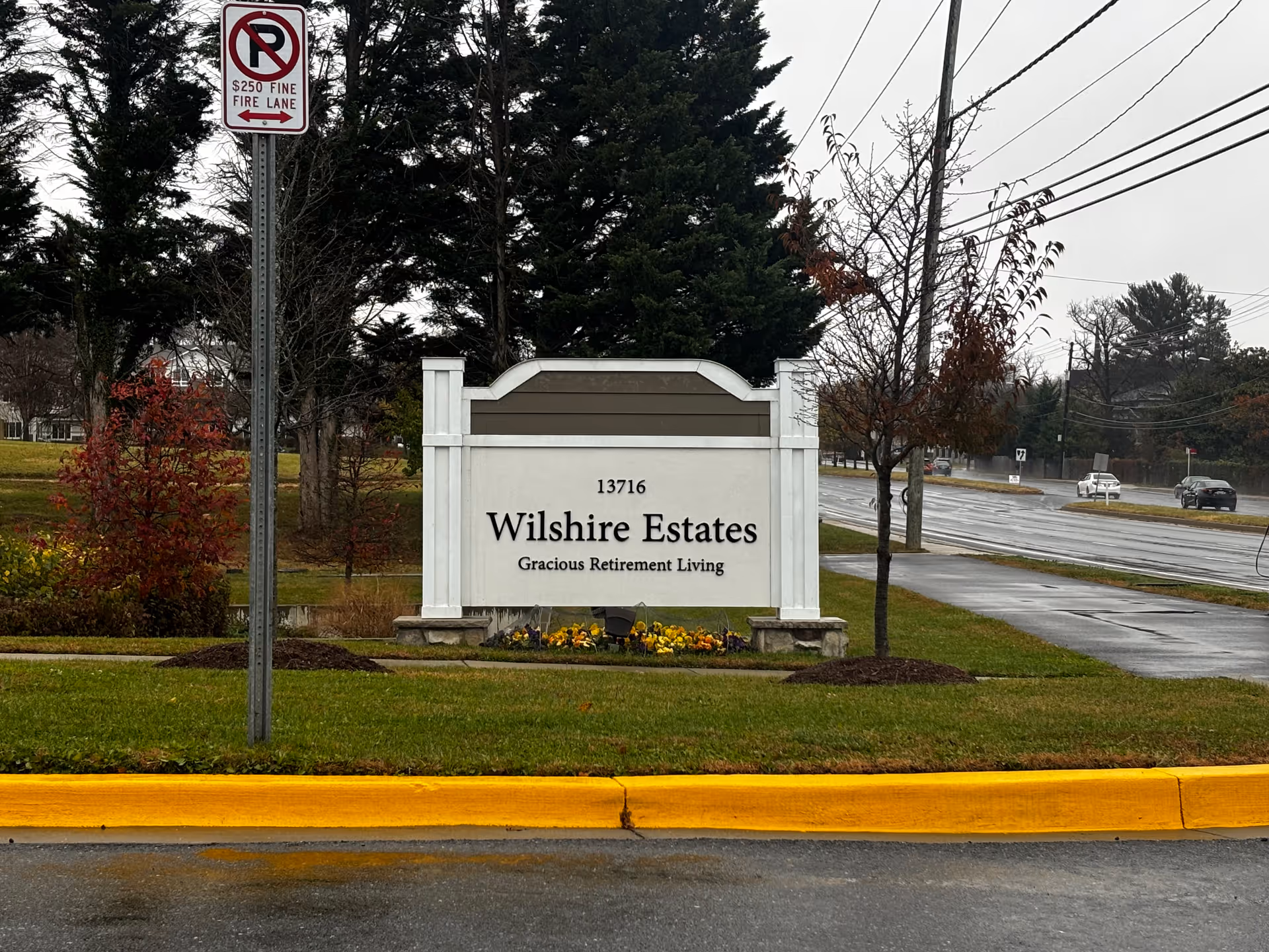 Outdoor view of a white and brown sign for Wilshire Estates Gracious Retirement Living, located on a grassy area next to a road with cars and trees in the background. A no parking fire lane sign is visible on a pole to the left of the sign.