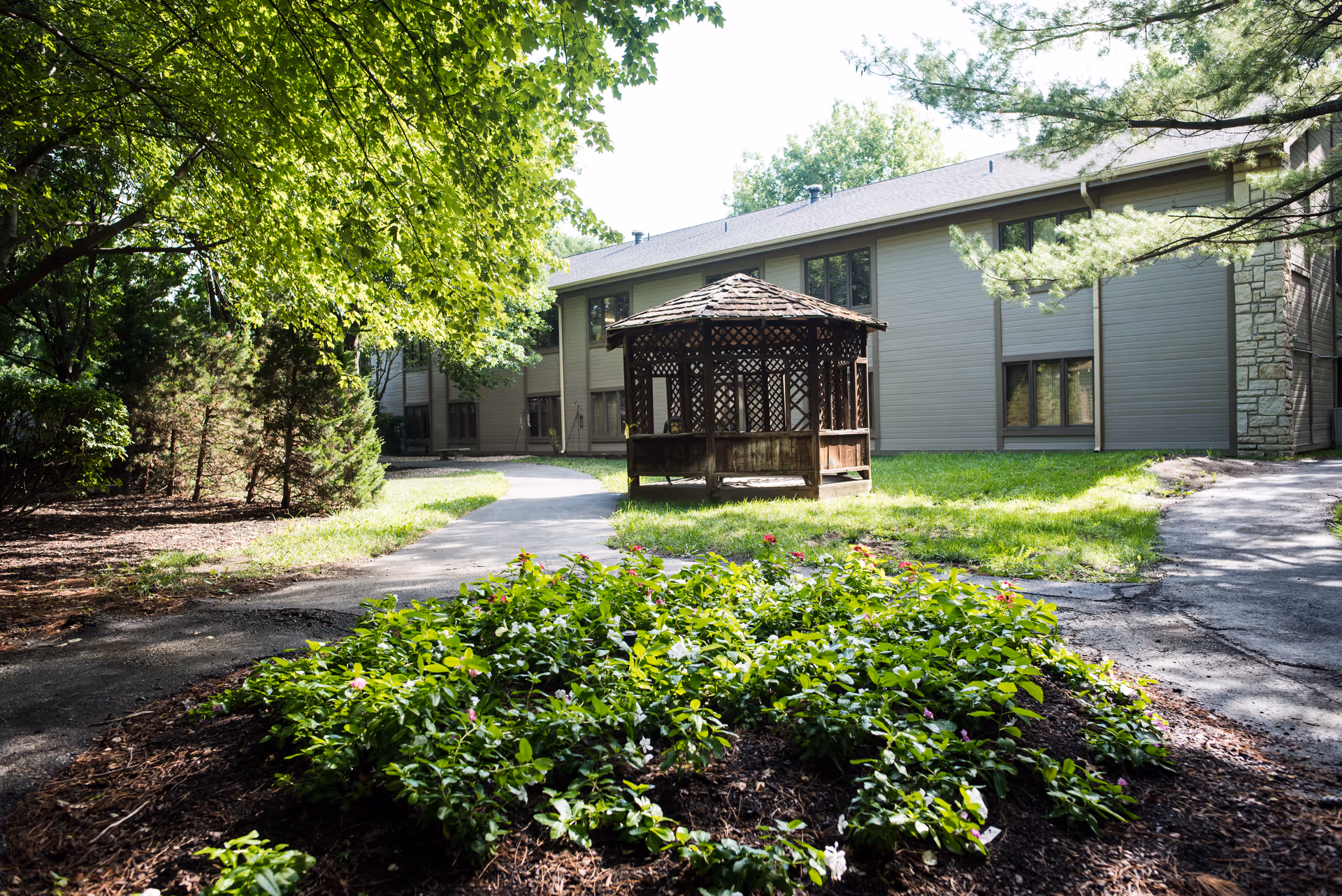 Outdoor garden area at Shawnee Gardens Healthcare and Rehabilitation Center featuring a wooden gazebo, surrounded by green grass, trees, and flowering plants with a two-story building in the background.