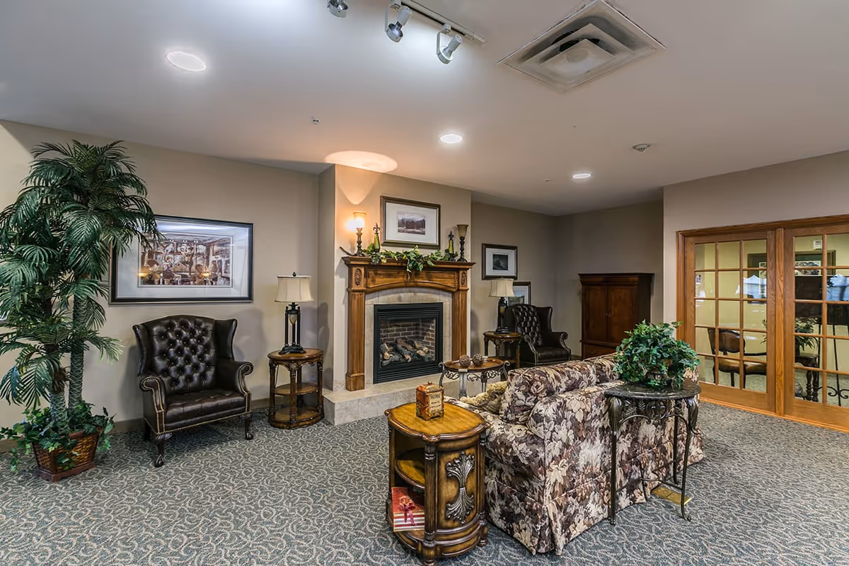 A cozy living room area featuring a floral-patterned sofa, two dark leather armchairs, a wooden fireplace with decorative items on the mantel, two side tables with lamps, a large potted plant, framed artwork on the walls, and glass-paneled double doors leading to another room.