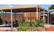Outdoor covered patio area with metal roof and several black metal chairs and tables. The patio is surrounded by a wooden fence and has a garden bed with various green plants and flowers in front of it. The sky is clear and blue.