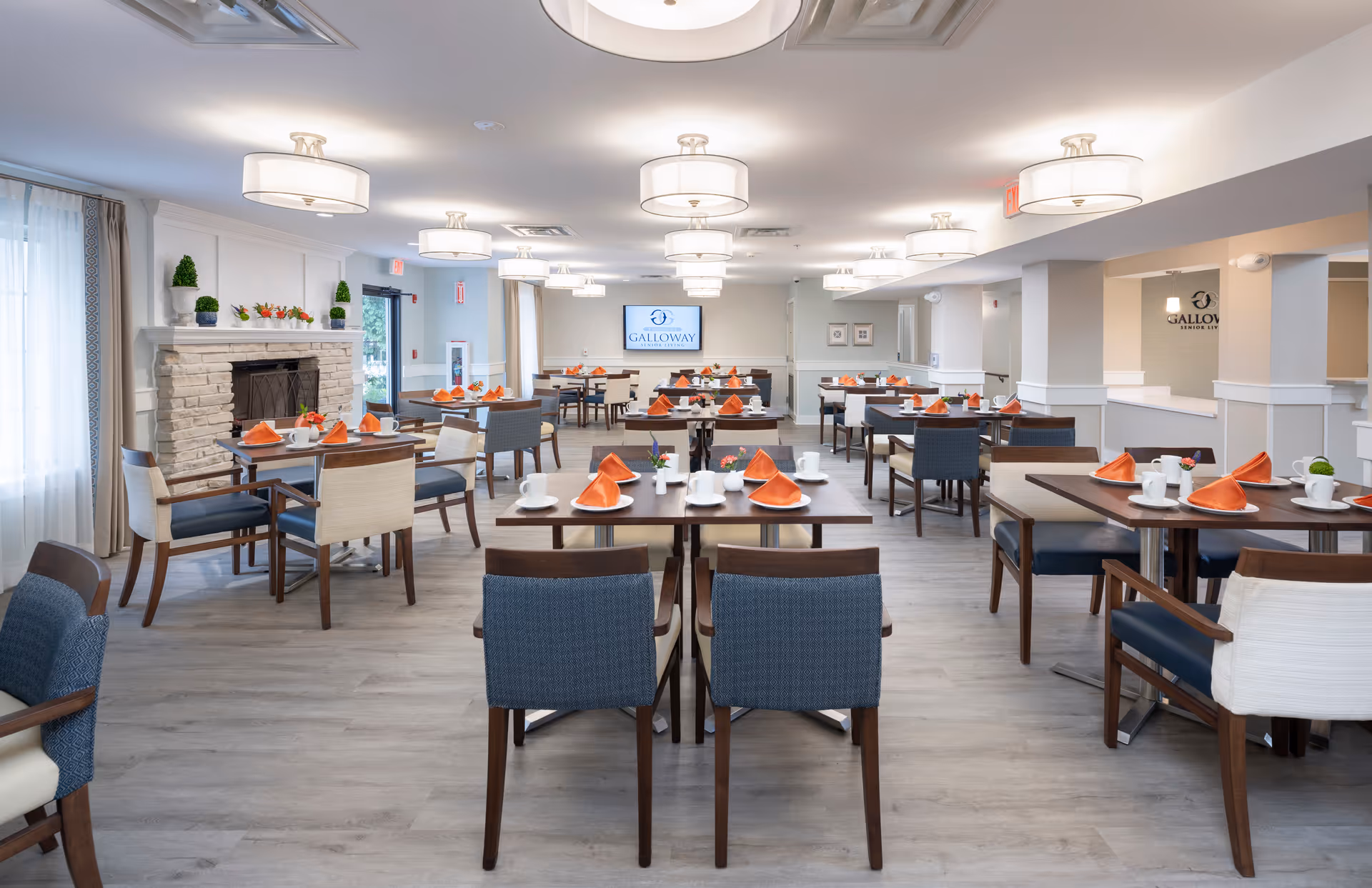A bright and spacious dining room in a senior living facility with multiple tables set with white cups, plates, and orange napkins. The room features modern light fixtures on the ceiling, a stone fireplace on the left side, and large windows with curtains allowing natural light to enter. The back wall has a screen displaying the Galloway Senior Living logo.
