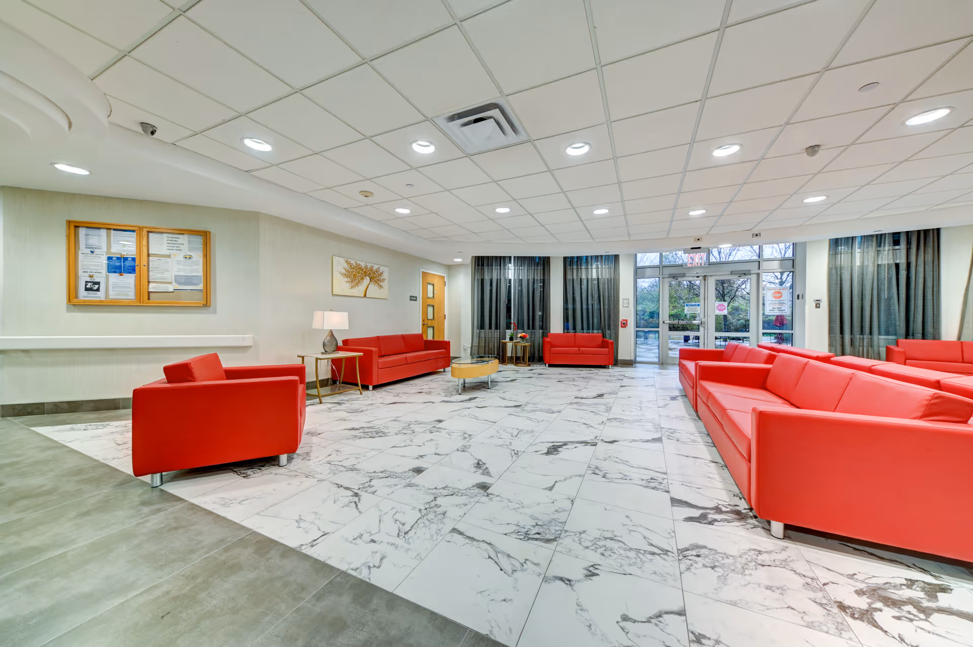 A spacious and well-lit lobby area with multiple red sofas arranged around a marble-tiled floor. The room has large windows with dark curtains, a glass entrance door, a bulletin board on the wall, a side table with a lamp, and a ceiling with recessed lighting.