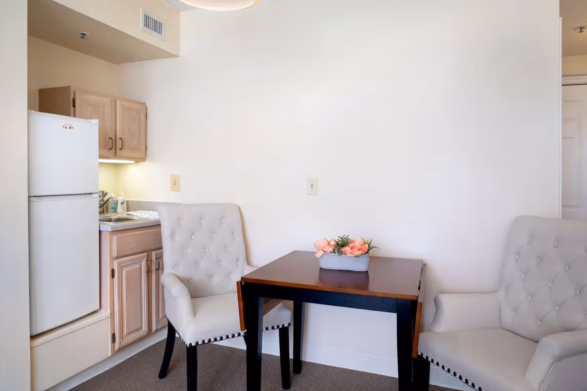 Small kitchenette area with a white refrigerator, light wood cabinets, and a stainless steel sink. In front of the kitchenette is a dark wood table with a small floral arrangement on top, flanked by two beige upholstered chairs with button tufting and nailhead trim. The walls are plain white and the floor is carpeted.
