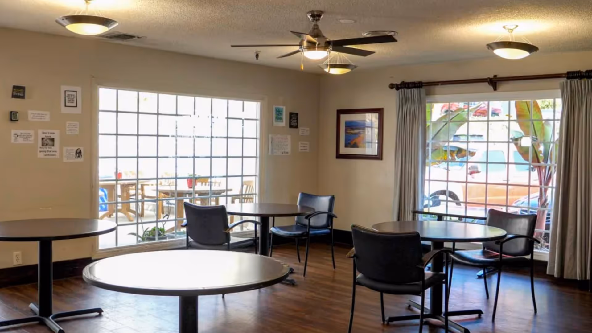 A bright room with round tables and chairs arranged for seating. Large windows with grid patterns let in natural light and show an outdoor patio area and parked vehicles outside. The room has wooden flooring, beige walls with framed pictures and small wall decorations, ceiling fans, and light fixtures.