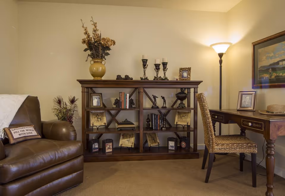 Cozy living room area with a leather armchair, wooden bookshelf filled with decor and a desk with a woven chair under warm lighting.