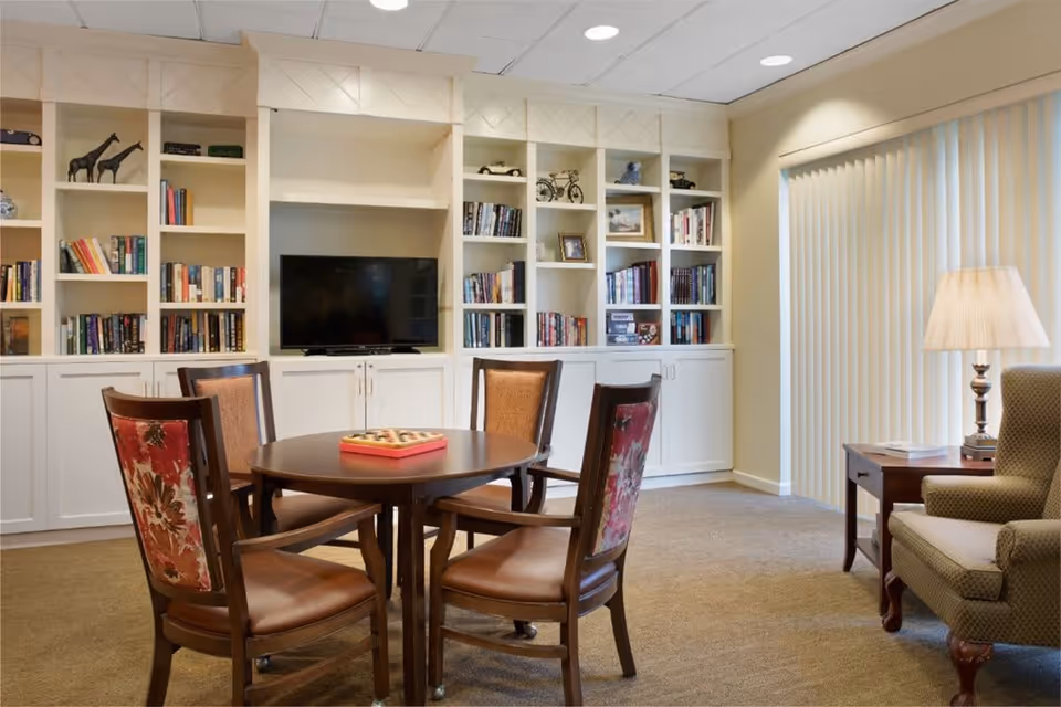 A cozy common area with a round wooden table surrounded by four chairs, each with floral upholstery on the backrest. Behind the table is a built-in white bookshelf filled with books, decorative items, and a flat-screen TV. To the right, there is a side table with a lamp and an upholstered armchair next to vertical blinds covering a window or sliding door.