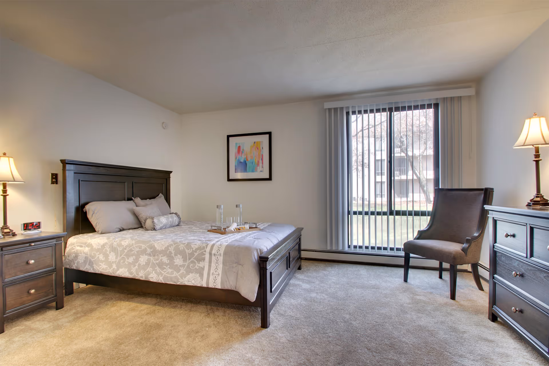 Bright furnished bedroom with a bed, nightstands and lamps, a chair and dresser beside a large window with vertical blinds.