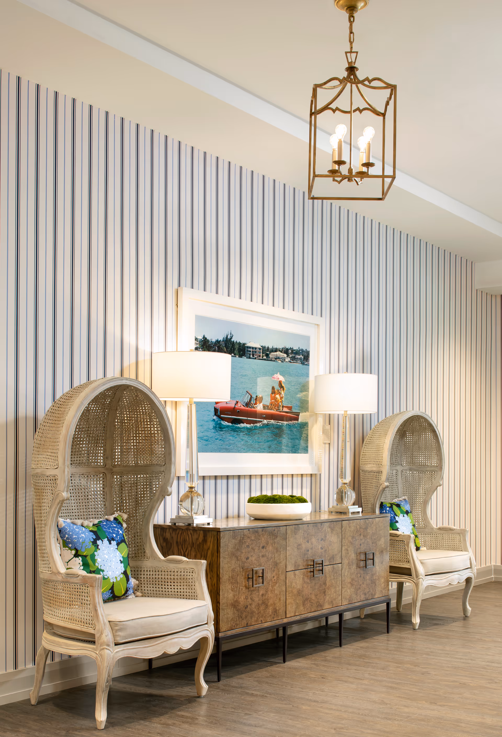 A cozy interior seating area with two high-backed wicker chairs featuring floral cushions, positioned on either side of a wooden sideboard with two table lamps and a decorative bowl. Above the sideboard hangs a framed picture of people in a red boat on a lake. The wall behind has vertical striped wallpaper and a hanging lantern-style light fixture on the ceiling.