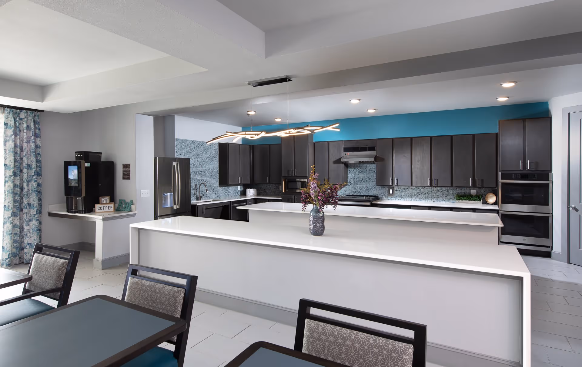 Modern kitchen area with dark wood cabinets, a large white island countertop with a vase of flowers, stainless steel appliances including a refrigerator, oven, and microwave, and a coffee station on the side. The backsplash is made of small blue tiles and the walls are painted gray and teal. Dining tables and chairs are partially visible in the foreground.
