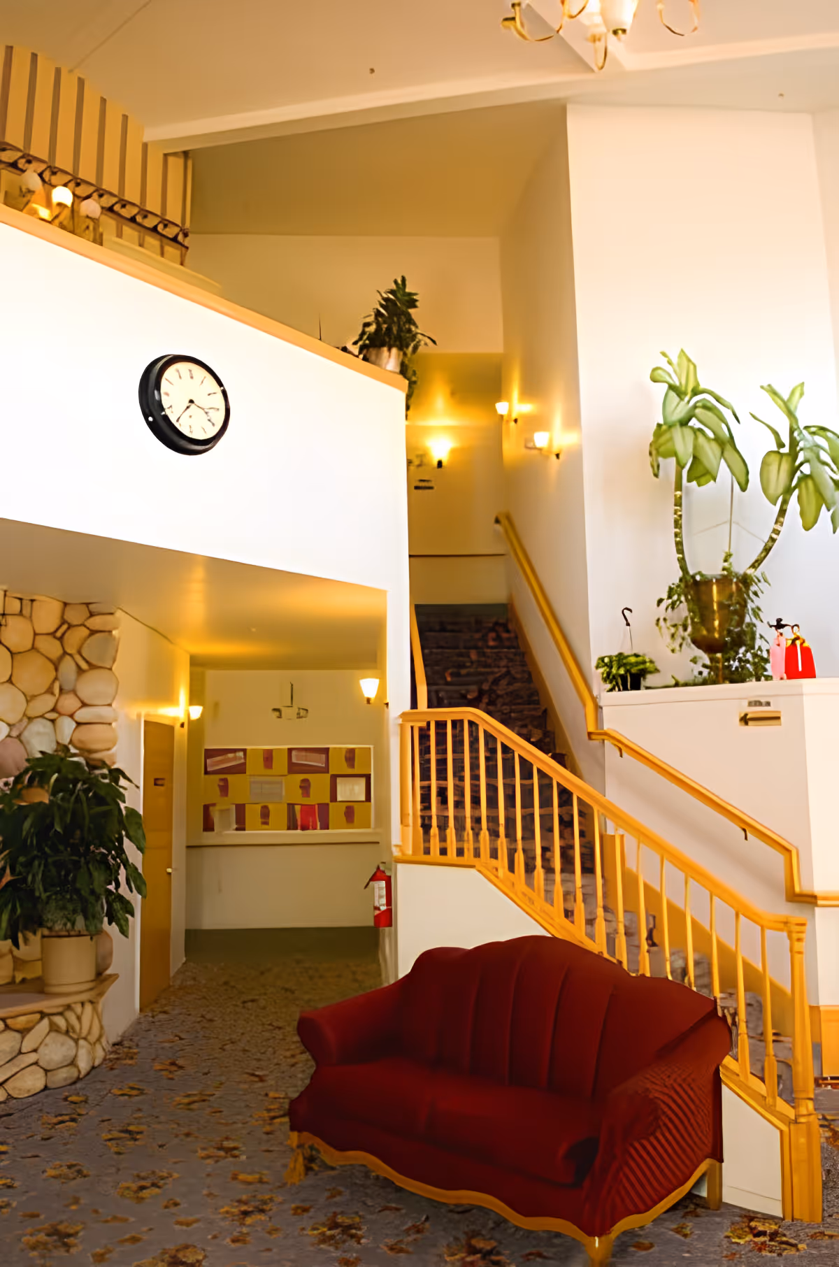 Lobby-style interior with a red upholstered sofa, staircase with wooden railing, wall clock, and potted plants.