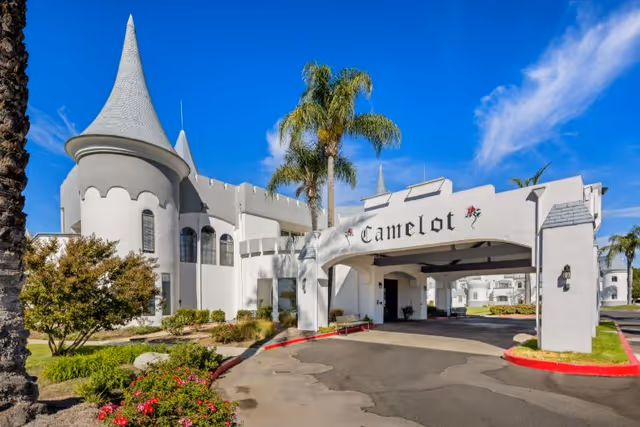 Front entrance of a white, castle-like senior living building with a drive-through canopy labeled "Camelot," palm trees, and a curved driveway.