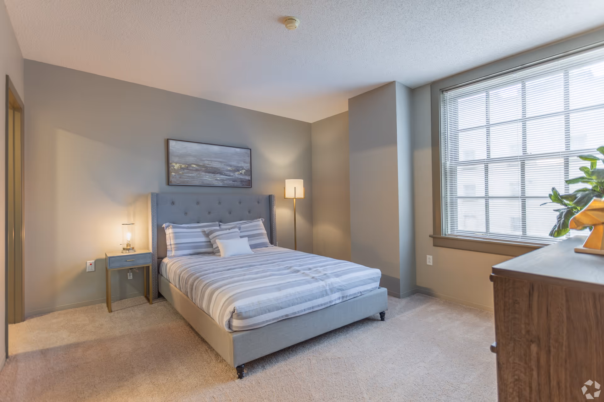 A neatly made bed with a gray upholstered headboard and striped bedding in a bedroom with beige walls and carpet. There is a small nightstand with a lamp on the left side of the bed and a tall floor lamp in the corner. A large window with blinds is on the right side, and a wooden dresser with a plant on top is partially visible. A framed abstract painting hangs above the bed.