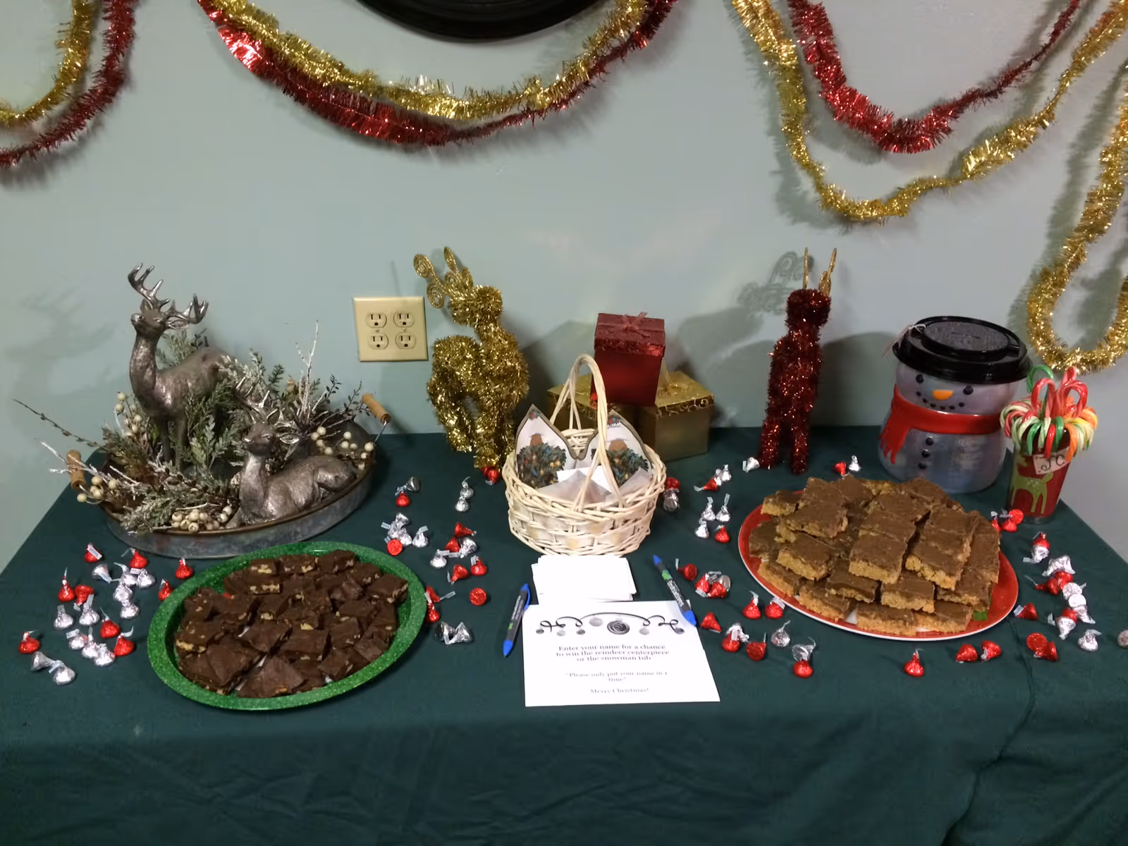 A festive holiday table decorated with tinsel garlands on the wall, two plates of brownies, a basket with pine cones, wrapped gift boxes, a snowman container, candy canes in a cup, and scattered Hershey's Kisses chocolates. There are also decorative reindeer figures and a sign inviting people to enter their name for a chance to win a prize.