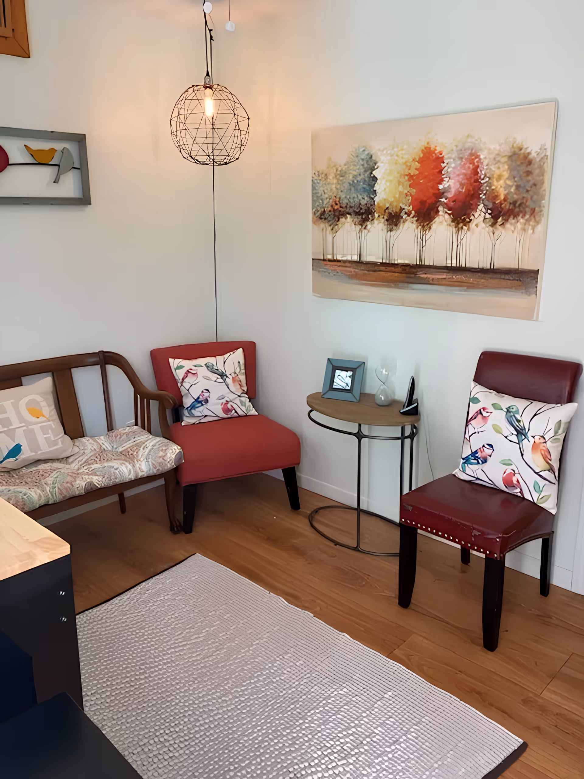 Small seating area with two chairs, a bench, a side table, a hanging wire pendant light, and a colorful tree painting on the wall.