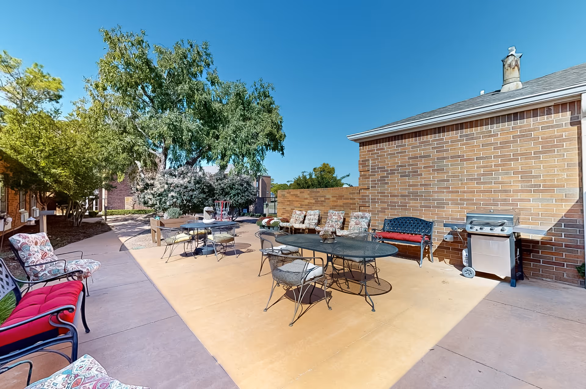 Outdoor patio area at Rivermont Assisted Living and Memory Care with several metal tables and chairs, cushioned benches, a barbecue grill, and trees providing shade under a clear blue sky.