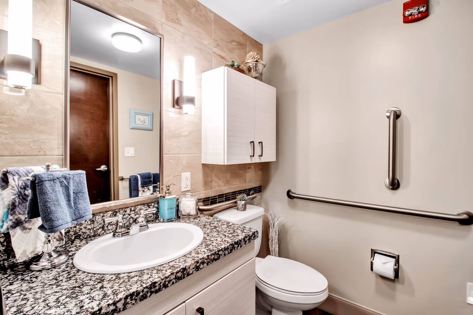 A clean and modern bathroom featuring a granite countertop with a white sink, a large mirror with two vertical light fixtures on either side, a white cabinet mounted on the wall above the toilet, and stainless steel grab bars on the wall next to the toilet. The bathroom has beige walls and a wooden door reflected in the mirror.