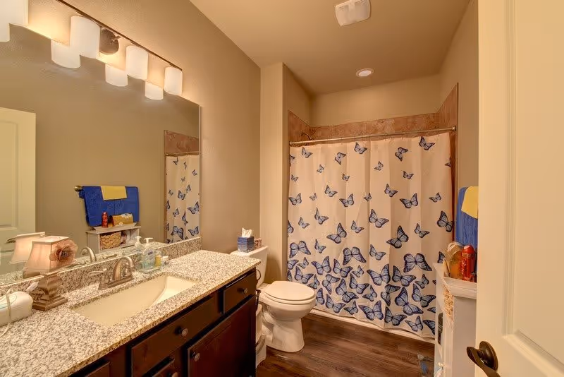 A bathroom with a granite countertop vanity featuring a sink and faucet, a large mirror with four light fixtures above it, a toilet, and a shower with a white curtain decorated with blue butterflies. The bathroom has beige walls and dark wood flooring. Towels and toiletries are visible on a small shelf and towel rack.
