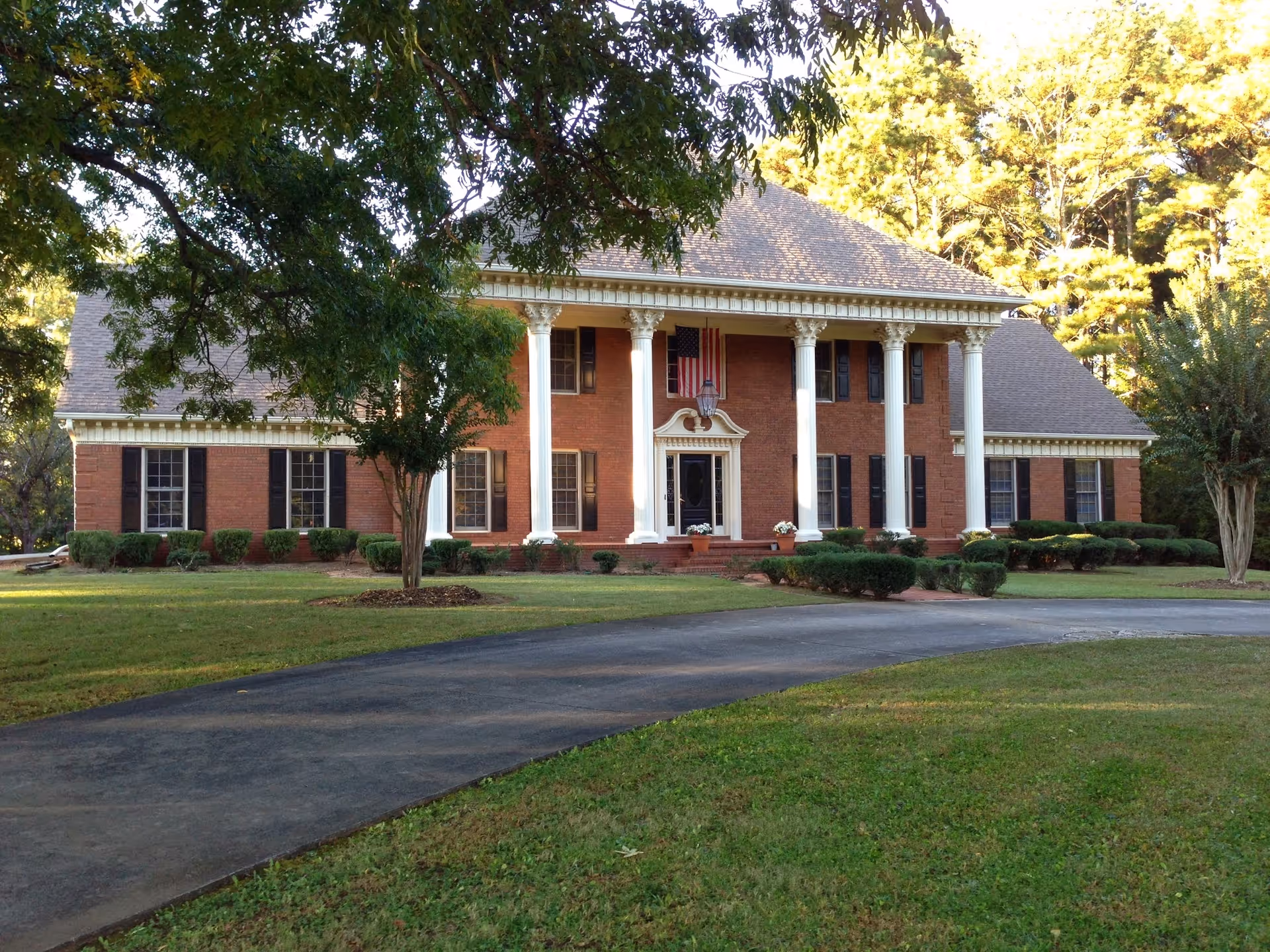 A large two-story brick building with white columns at the entrance, black shutters on the windows, and an American flag hanging above the front door. The building is surrounded by a well-maintained lawn with bushes and trees, and a curved driveway leads up to the entrance.
