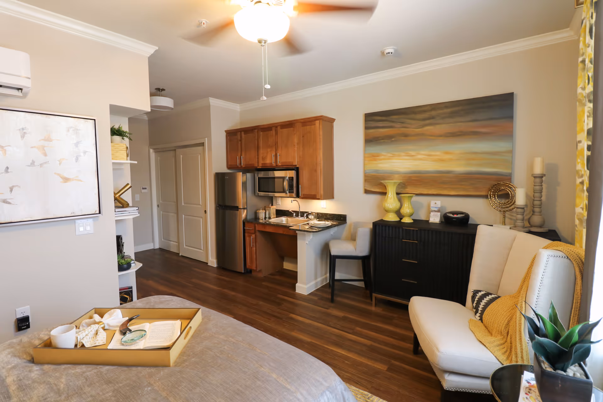 A cozy assisted living apartment interior featuring a small kitchenette with wooden cabinets, a stainless steel refrigerator, and a microwave. The room includes a comfortable beige armchair with a yellow throw blanket, a black dresser with decorative vases and candles, and a bed with a tray holding a cup, napkin, and an open book. The walls are adorned with a large painting and a framed artwork, and the floor is covered with wood-style flooring.