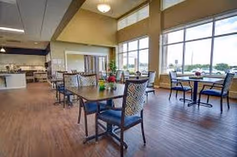 Bright dining area with tables, chairs, and large windows in a senior living facility.