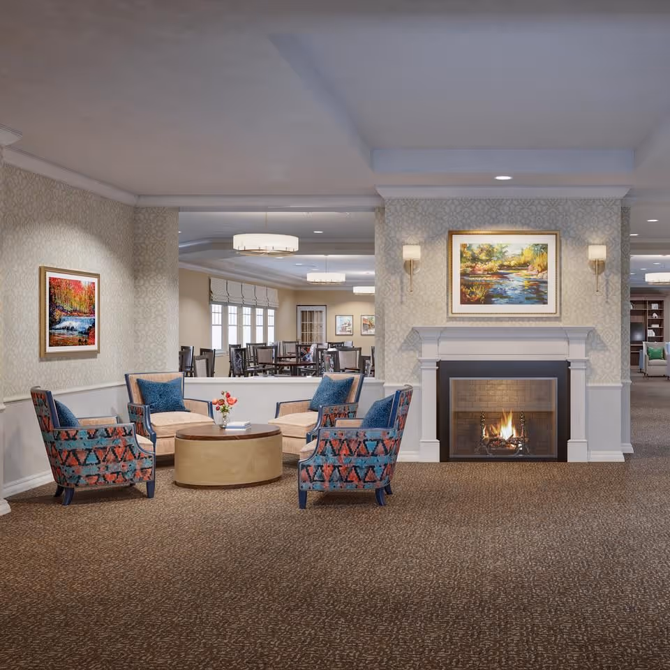 A cozy common area with four patterned armchairs and two beige chairs arranged around a round wooden coffee table with a small flower vase. The room features a lit fireplace with a colorful landscape painting above it, soft wall sconces, and patterned wallpaper. In the background, there is a dining area with multiple tables and chairs, and additional artwork on the walls.