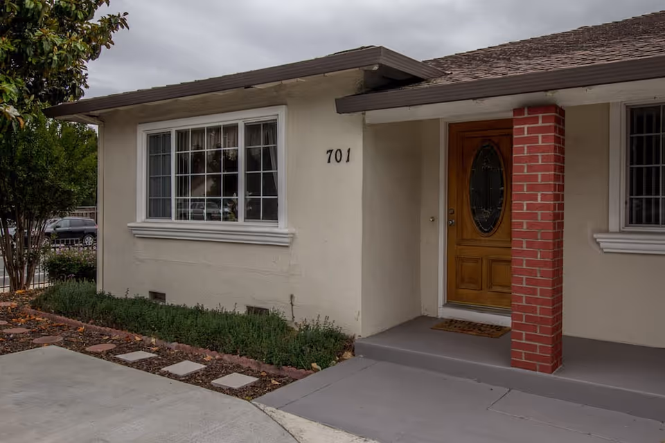 Exterior view of a single-story building with beige walls, a brown shingled roof, and a wooden front door with an oval glass panel. The building has a red brick column supporting a small porch roof, a large window with white trim, and the number 701 displayed on the wall. There are some shrubs and a tree near the building, and a concrete driveway in the foreground.