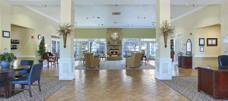 Spacious and well-lit common area with a stone fireplace centered on the far wall, surrounded by armchairs and tables. The room has large windows letting in natural light, light-colored walls, and a mix of tiled and carpeted flooring. Decorative plants and framed pictures adorn the walls, creating a welcoming atmosphere.