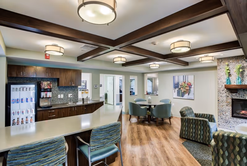 A modern common area in a senior living facility featuring a long countertop with bar stools, a beverage refrigerator stocked with drinks, wooden cabinets, and a coffee machine. The room has wood-beamed ceilings, a round table with chairs, patterned armchairs, a stone fireplace with decorative vases, and wood flooring.