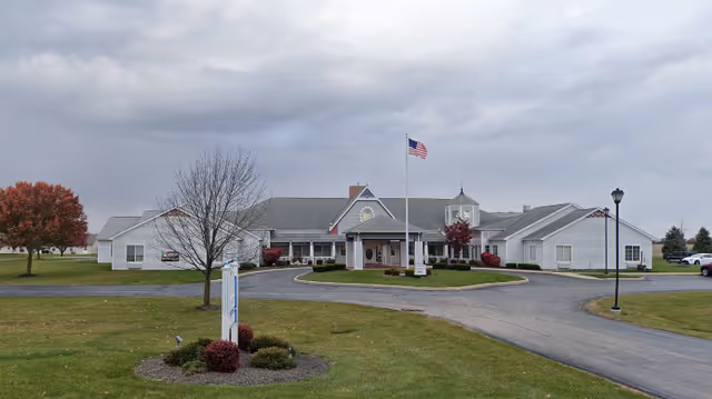 Front exterior view of a single-story senior living facility building with a circular driveway, an American flag on a flagpole, and landscaped grounds with trees and bushes under a cloudy sky.