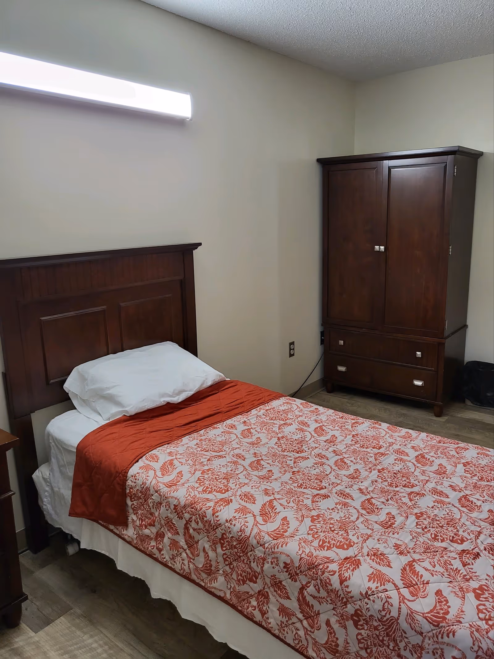 A small bedroom with a single bed featuring a wooden headboard, white pillow, white sheets, and a red and white floral patterned blanket. There is a wooden armoire with drawers against the wall and a small wooden nightstand partially visible. The walls are plain and light-colored, and the floor has a wood-like finish.