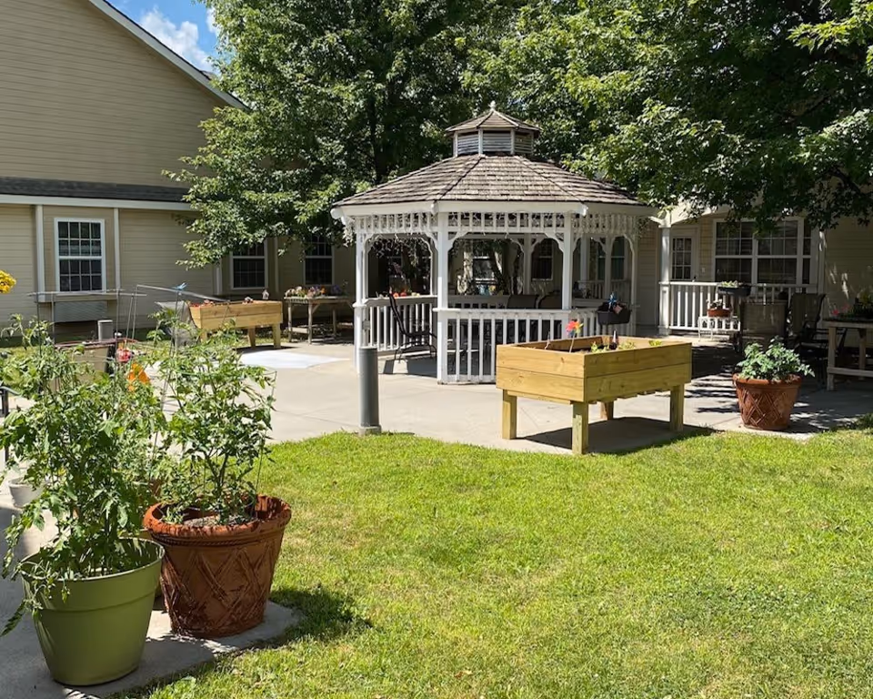 Outdoor courtyard area at Marquis Place of Elkhorn featuring a white wooden gazebo with a shingled roof, surrounded by green grass, potted plants, and raised garden beds. The background shows a beige building with windows and a shaded patio area with chairs and tables.