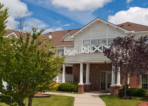 Exterior view of a two-story senior living facility building with a brick and white siding facade, a balcony with white railings, and a pathway leading to the entrance. There are green trees and shrubs around the building under a partly cloudy blue sky.