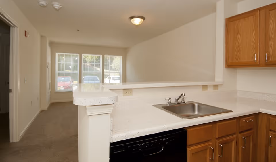 Interior view of a kitchen area with wooden cabinets, a stainless steel sink, and a white speckled countertop. The kitchen opens into a living room area with large windows letting in natural light. The walls are painted white and the floor is carpeted.