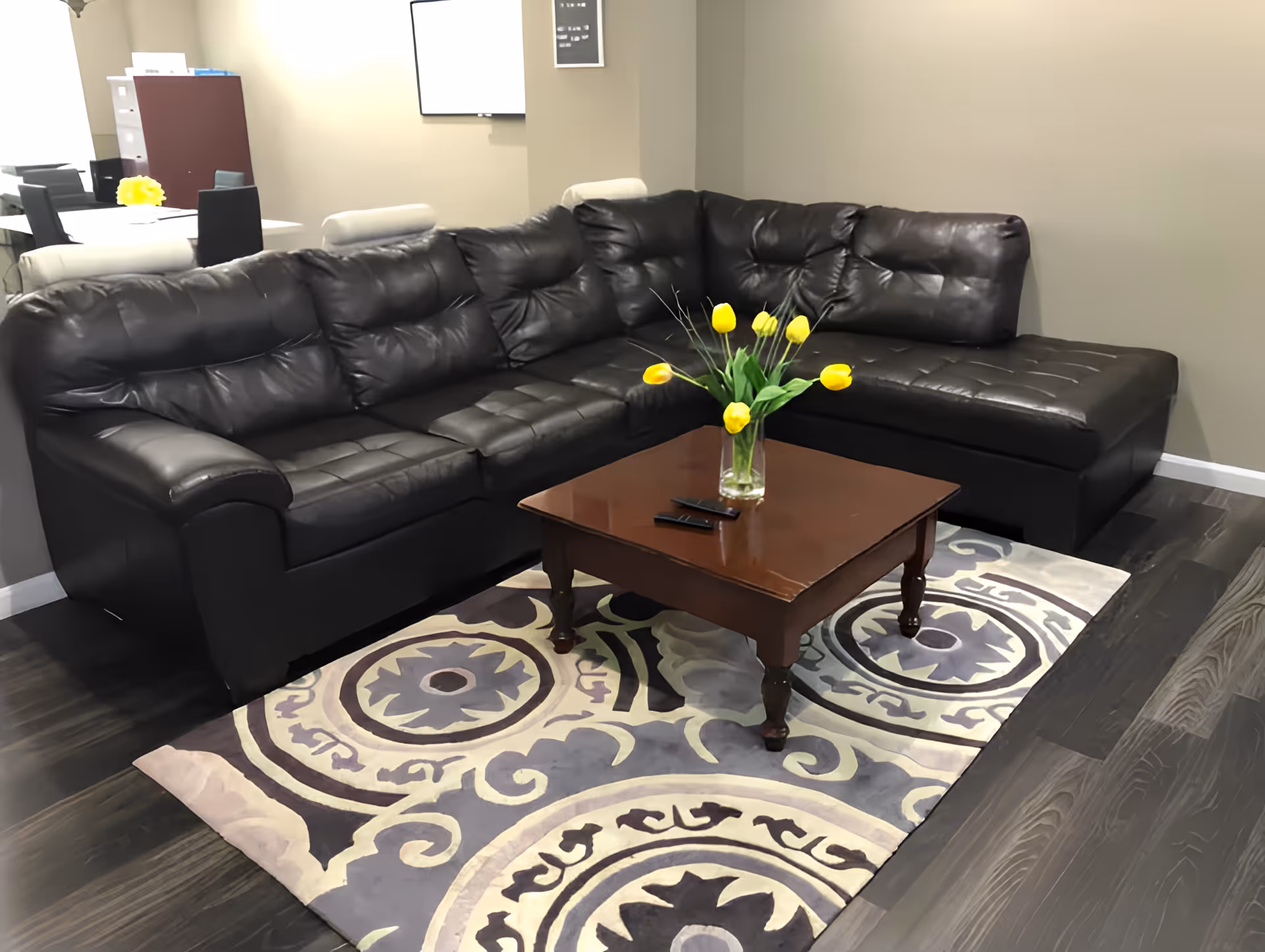 A cozy living room area with a large black leather sectional sofa, a wooden coffee table with a glass vase of yellow tulips, and a patterned area rug on dark hardwood flooring. In the background, there is a dining table with chairs and a cabinet against the wall.