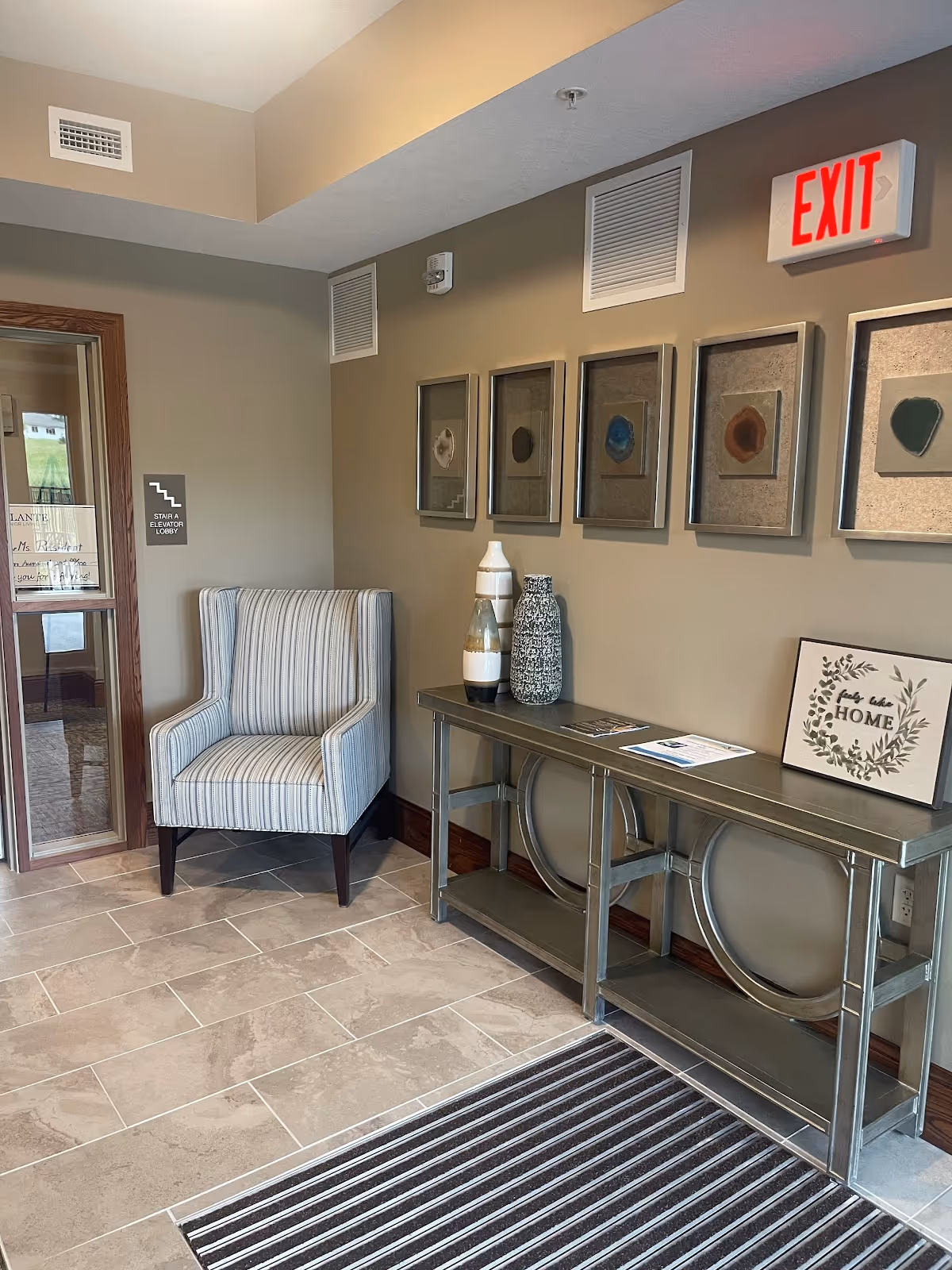 Entryway lobby with a striped armchair, a console table holding decorative vases and framed art on the wall beneath an EXIT sign.
