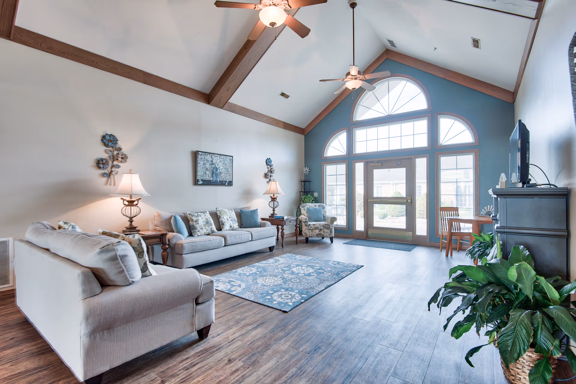 A bright and spacious living room with vaulted ceilings and wooden beams. The room features two beige sofas with patterned cushions, a matching armchair, two side tables with lamps, a blue patterned rug, and a large windowed door with an arched window above it letting in natural light. There is a TV on a dark wooden stand and some green plants in the corner.