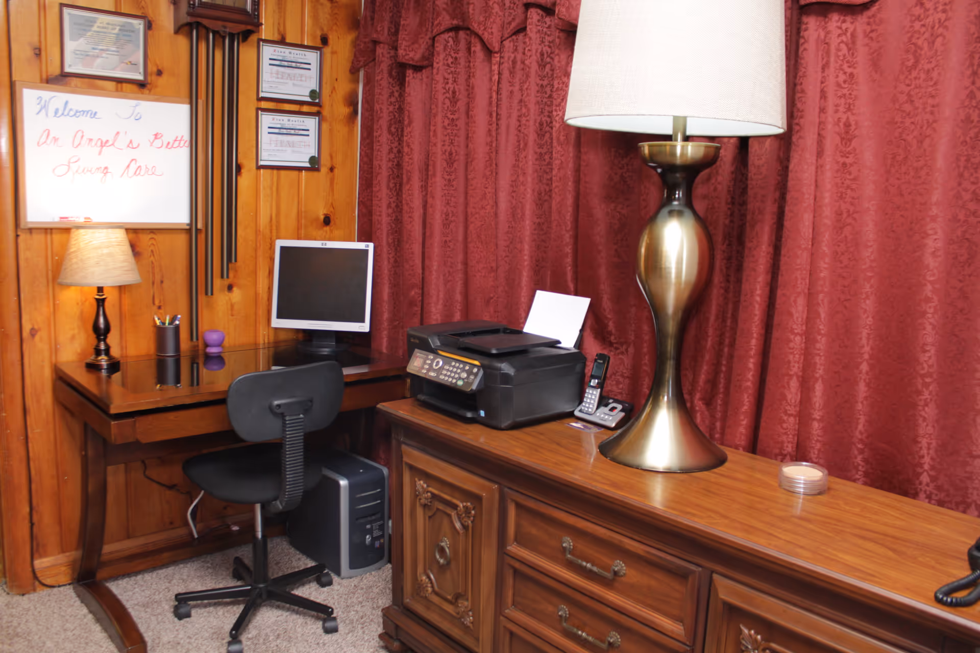 Wood-paneled interior room with a desk area, computer and printer, lamps, and red curtains.