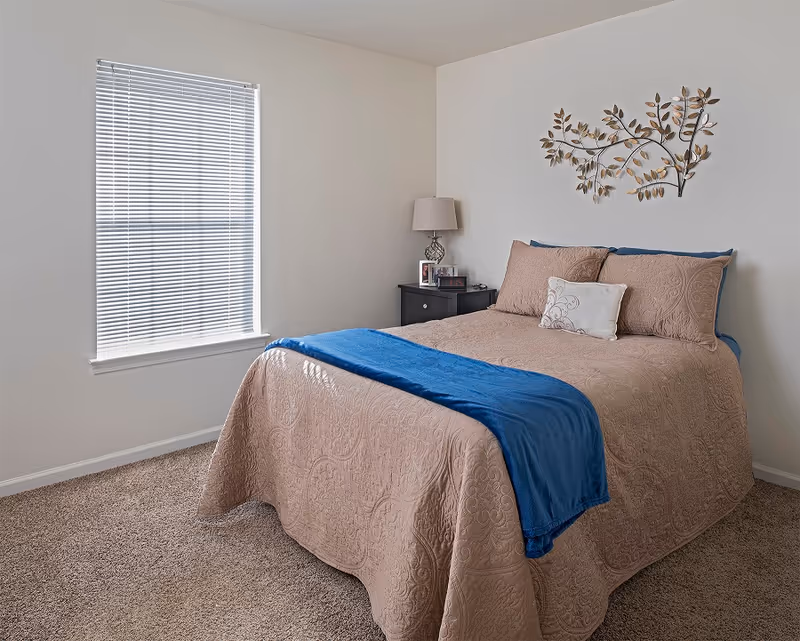 A neatly made bed with beige quilted bedding and a blue throw blanket at the foot. There are two large beige pillows and one smaller decorative pillow. Next to the bed is a black nightstand with a lamp, framed photos, and a small clock. A decorative metal wall art piece shaped like branches with leaves hangs above the bed. The room has beige carpet and a window with closed blinds letting in natural light.