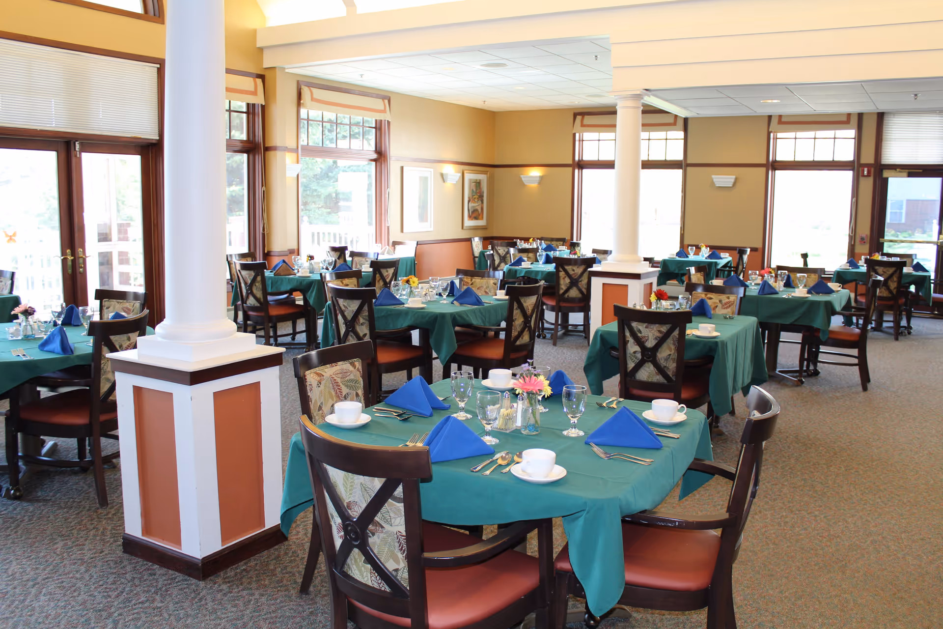 A bright and spacious dining room with multiple round tables covered in green tablecloths and set with blue folded napkins, white cups and saucers, glassware, and silverware. The room features large windows allowing natural light to fill the space, beige walls with framed artwork, and carpeted floors. There are wooden chairs with patterned backs around each table, and white columns supporting the ceiling.