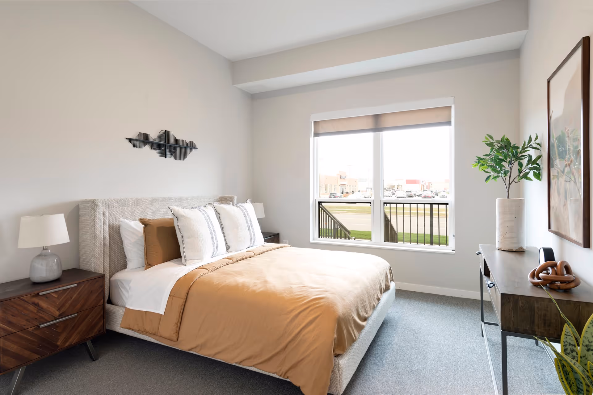 A bright bedroom with a large bed featuring tan and white bedding, two bedside tables with lamps, a decorative wall piece above the headboard, a window with a view of a parking lot outside, and a console table with a plant and decorative items.