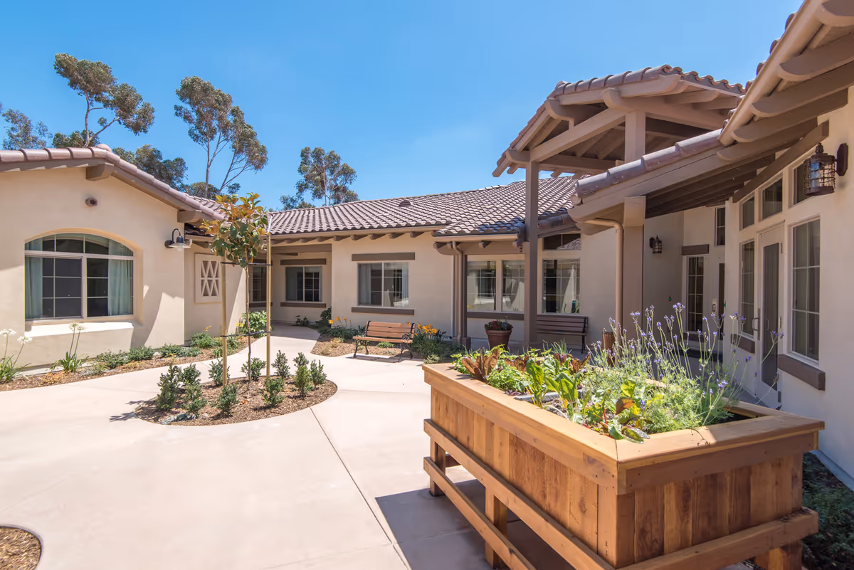 Outdoor courtyard area of a senior living facility with beige stucco buildings, tiled roofs, a wooden planter box with various plants, benches, and small landscaped garden beds under a clear blue sky.