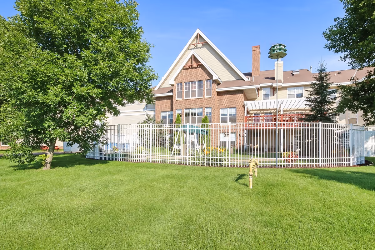 A brick-and-siding senior living building behind a white fence with a green lawn, trees, and a tall birdhouse under a clear blue sky.