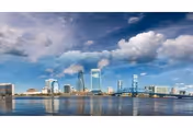 City skyline with tall buildings and a bridge over a body of water under a partly cloudy sky.
