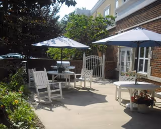 Sunlit outdoor patio with white tables and chairs, blue umbrellas, potted plants, and a brick building facade.