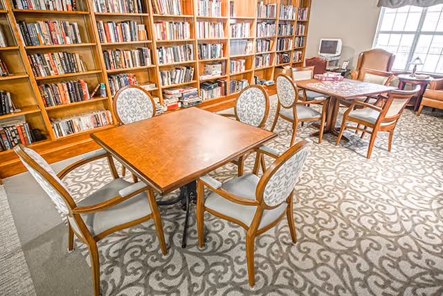 A cozy library or reading room with wooden bookshelves filled with books along the wall. In the foreground, there is a wooden table surrounded by six upholstered chairs with patterned fabric. In the background, another table with chairs and a small TV on a stand near a window with natural light coming in. The room has a patterned carpet and a comfortable, inviting atmosphere.