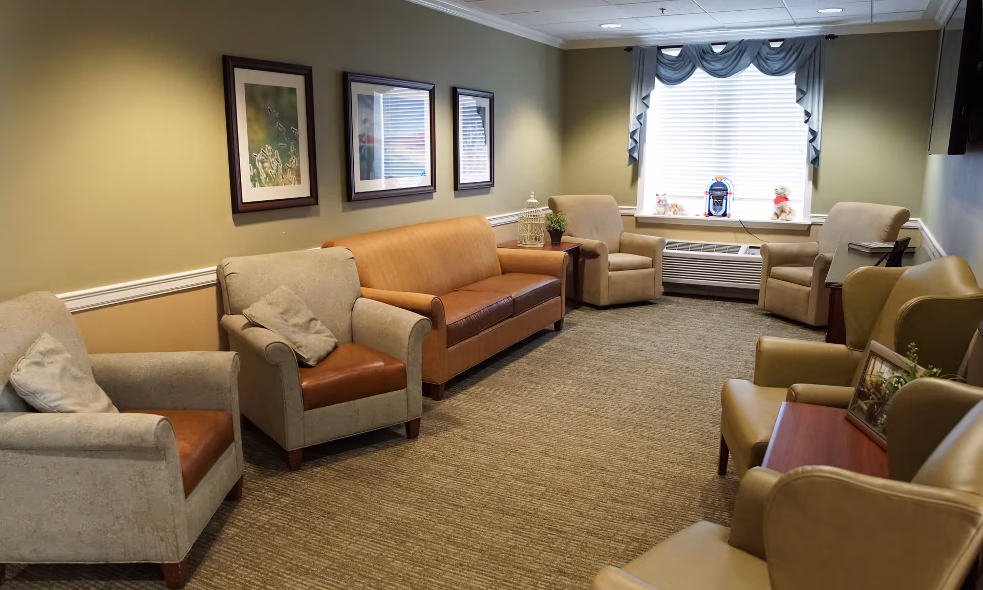 A cozy living room with beige and brown armchairs and a brown leather sofa arranged around the room. There are three framed pictures on the wall, a window with blinds and a blue valance, and a small jukebox and stuffed animals on the windowsill. The room has carpeted flooring and soft lighting.