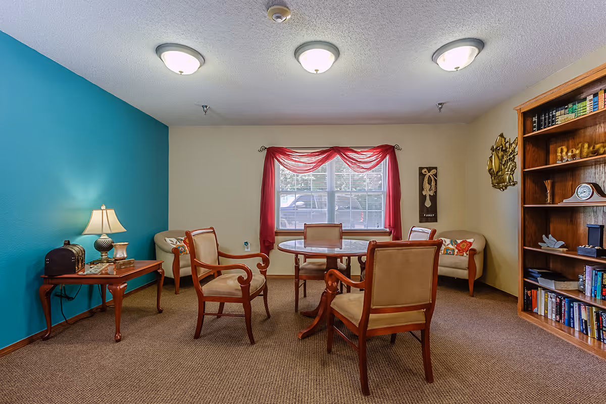A cozy common room with a round wooden table surrounded by four cushioned wooden chairs. The room has a teal accent wall on the left, beige walls elsewhere, and a window with red sheer curtains letting in natural light. There are two beige armchairs with colorful pillows near the window, a wooden side table with a lamp and decorative items, and a wooden bookshelf filled with books and decor on the right.