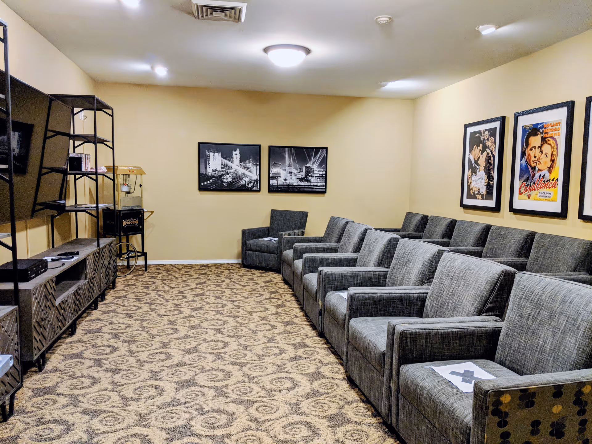A cozy media room with a row of gray upholstered armchairs facing a large flat-screen TV mounted on the wall. The room has beige walls adorned with framed black and white cityscape photos and classic movie posters. There is a patterned carpet on the floor and a popcorn machine in the corner.