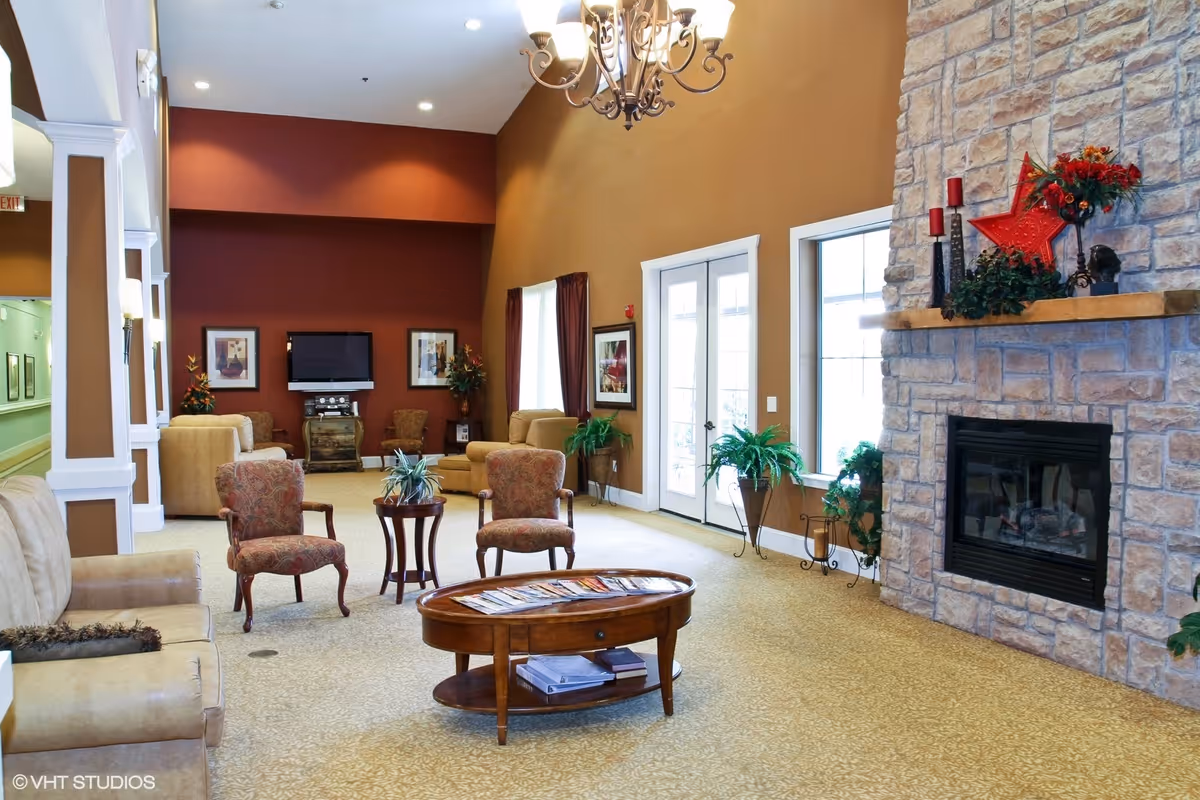 A spacious living room area with beige and patterned armchairs, a wooden coffee table with magazines, a stone fireplace decorated with red candles and a red star, and a wall-mounted TV. The room has warm brown walls, large windows with curtains, and a chandelier hanging from the ceiling.